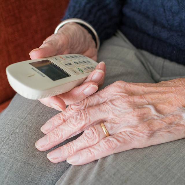 Die Hände einer älteren Person liegen im Schoß und halten ein Telefon