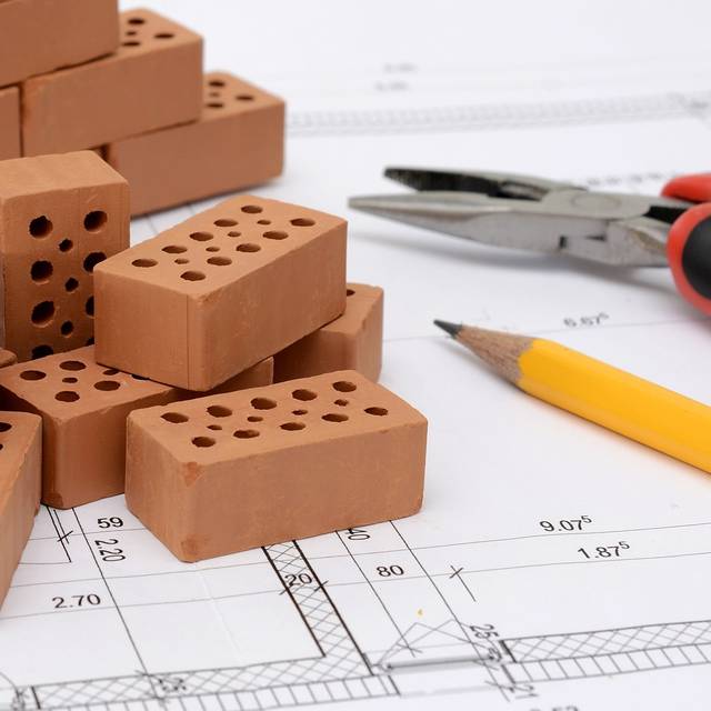 Auf einem Bausplan liegt ein Stift und eine Zange. Daneben rote Ziegelsteine.