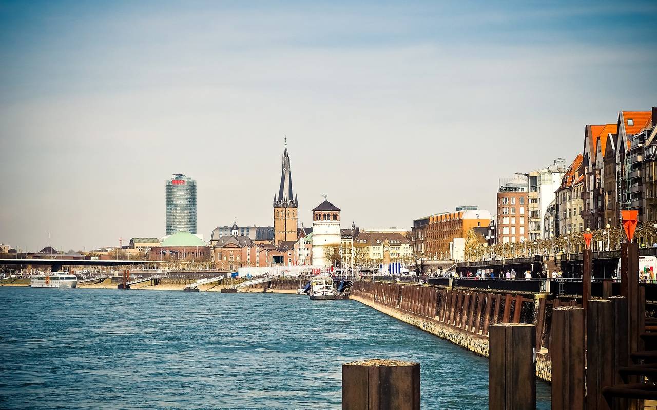 Ausblick auf die Düsseldorfer Rheinuferpromenade mit dem Schlossturm und der Basilika St. Lambertus im Hintergrund. Im linken Vordergrund ist der Rhein zu sehen.