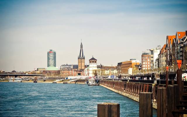 Ausblick auf die Düsseldorfer Rheinuferpromenade mit dem Schlossturm und der Basilika St. Lambertus im Hintergrund. Im linken Vordergrund ist der Rhein zu sehen.