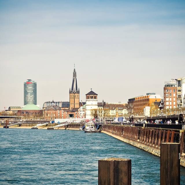 Ausblick auf die Düsseldorfer Rheinuferpromenade mit dem Schlossturm und der Basilika St. Lambertus im Hintergrund. Im linken Vordergrund ist der Rhein zu sehen.