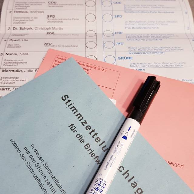 Die Briefwahlunterlagen - ein roter und ein blauer Umschlag liegen auf einem weißen Wahlzettel. Ganz oben liegt eins Stift zum wählen.
