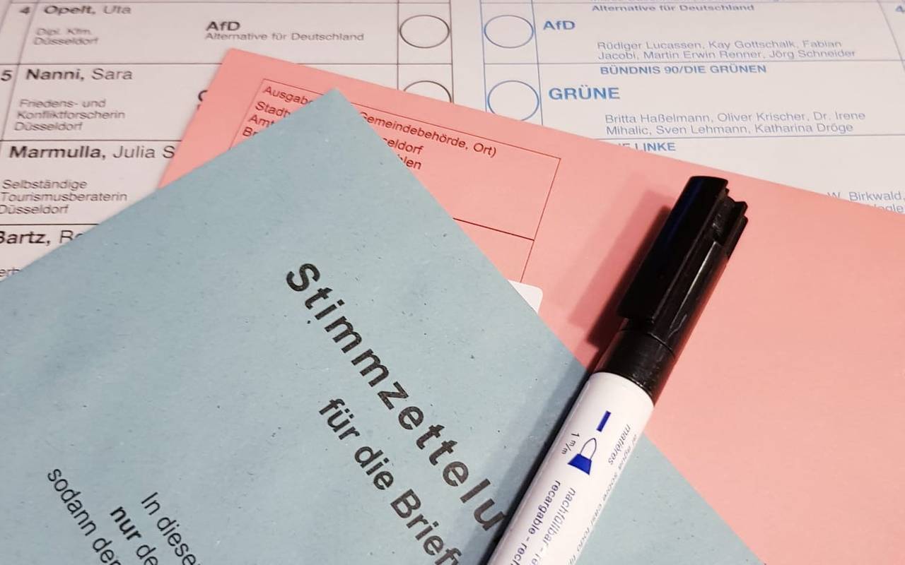 Die Briefwahlunterlagen - ein roter und ein blauer Umschlag liegen auf einem weißen Wahlzettel. Ganz oben liegt eins Stift zum wählen.
