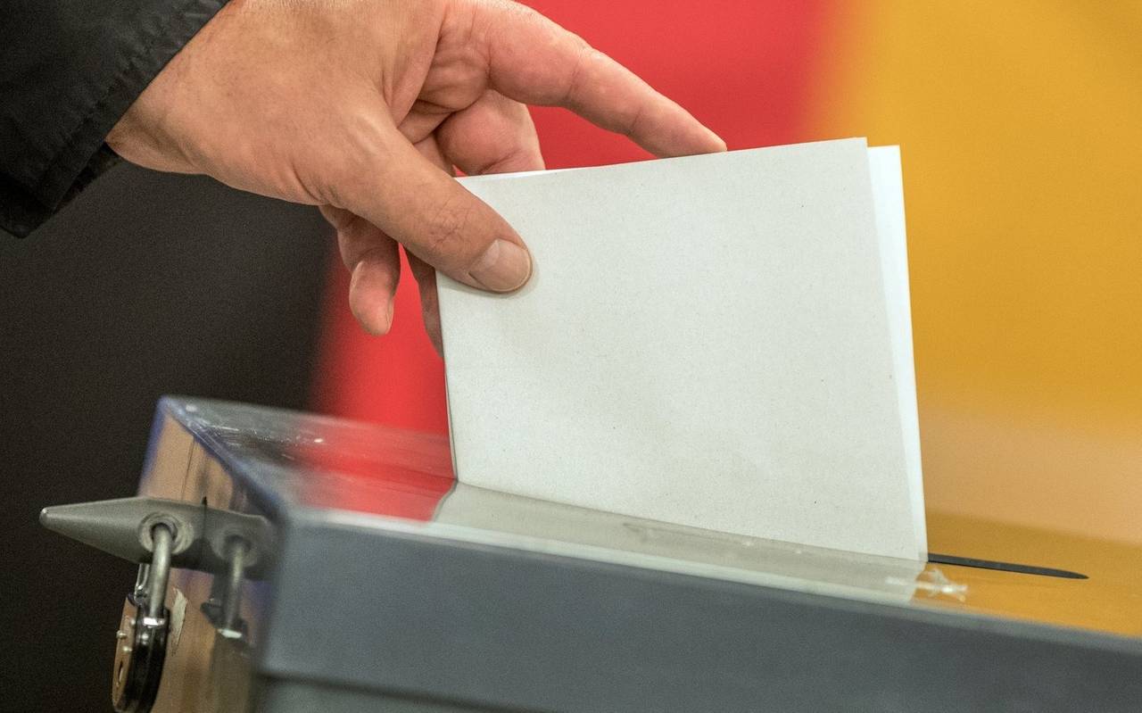 Ein Wahlzettel wird in eine Wahlurne eingeworfen. Im Hintergrund ist eine Deutschland-Fahne zu erkennen.