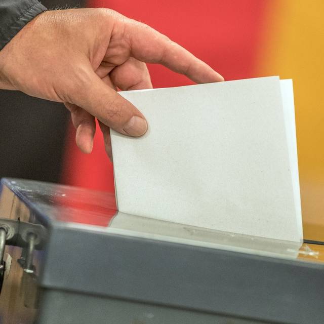 Ein Wahlzettel wird in eine Wahlurne eingeworfen. Im Hintergrund ist eine Deutschland-Fahne zu erkennen.