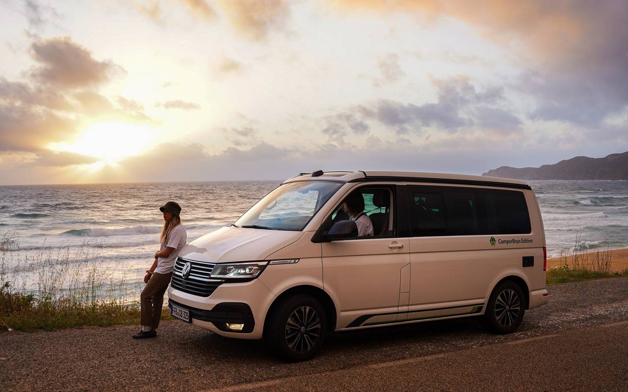 Ein Camper steht direkt am Meer. Die Sonne geht unter. Ein Mann lehnt entspannt am Wagen.
