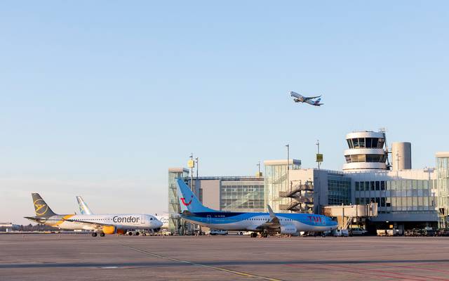 Eine Aufnahme vom Rollfeld des Düsseldorfer Flughafens. Zwei Flugzeuge stehen geparkt vor dem Flughafengebäude. Ein Flugzeug befindet sich in der Luft und startet gerade.