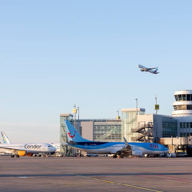 Eine Aufnahme vom Rollfeld des Düsseldorfer Flughafens. Zwei Flugzeuge stehen geparkt vor dem Flughafengebäude. Ein Flugzeug befindet sich in der Luft und startet gerade.