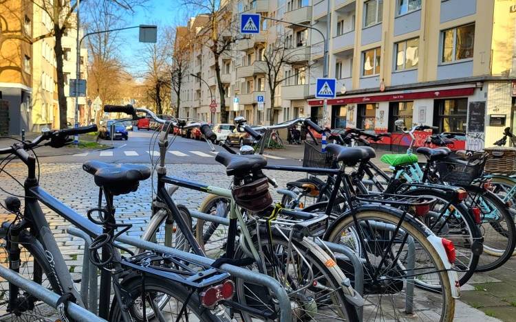 Mehrere Fahrräder stehen an Fahrradständern an einer Straße. Im Hintergrund sind Häuserfronten und ein Fußgängerüberweg zu sehen.