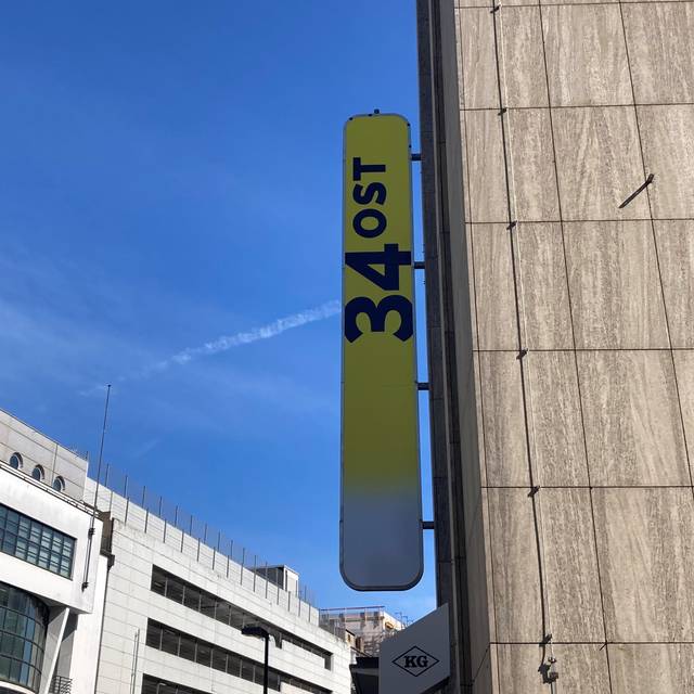 Ein gelbes Schild hängt draußen an einem grauen Gebäude. Darauf steht "34Ost".