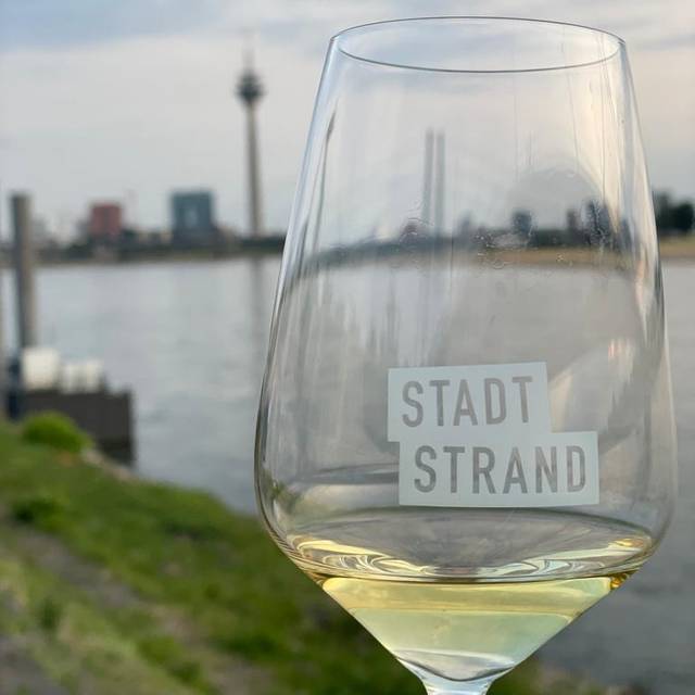 Ein Weinglas mit Weißwein und der Aufschrift "Stadtstrand", im Hintergrund ist die Düsseldorfer Skyline zu sehen