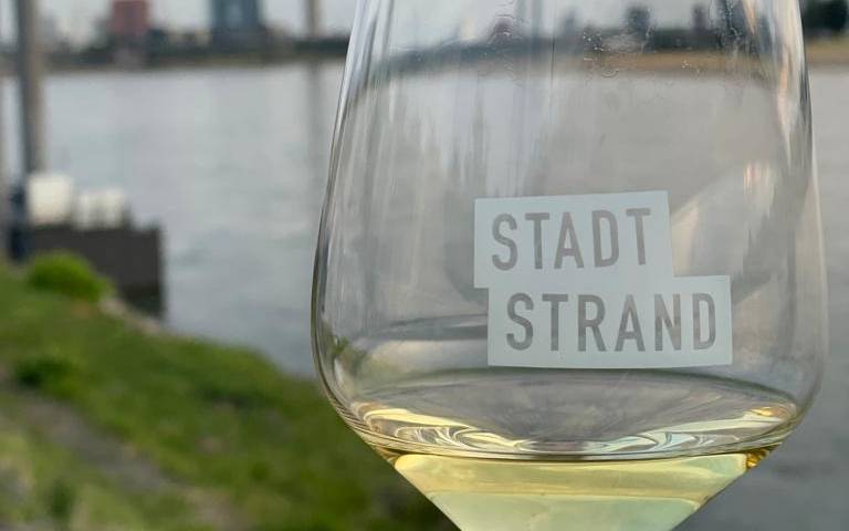 Ein Weinglas mit Weißwein und der Aufschrift "Stadtstrand", im Hintergrund ist die Düsseldorfer Skyline zu sehen