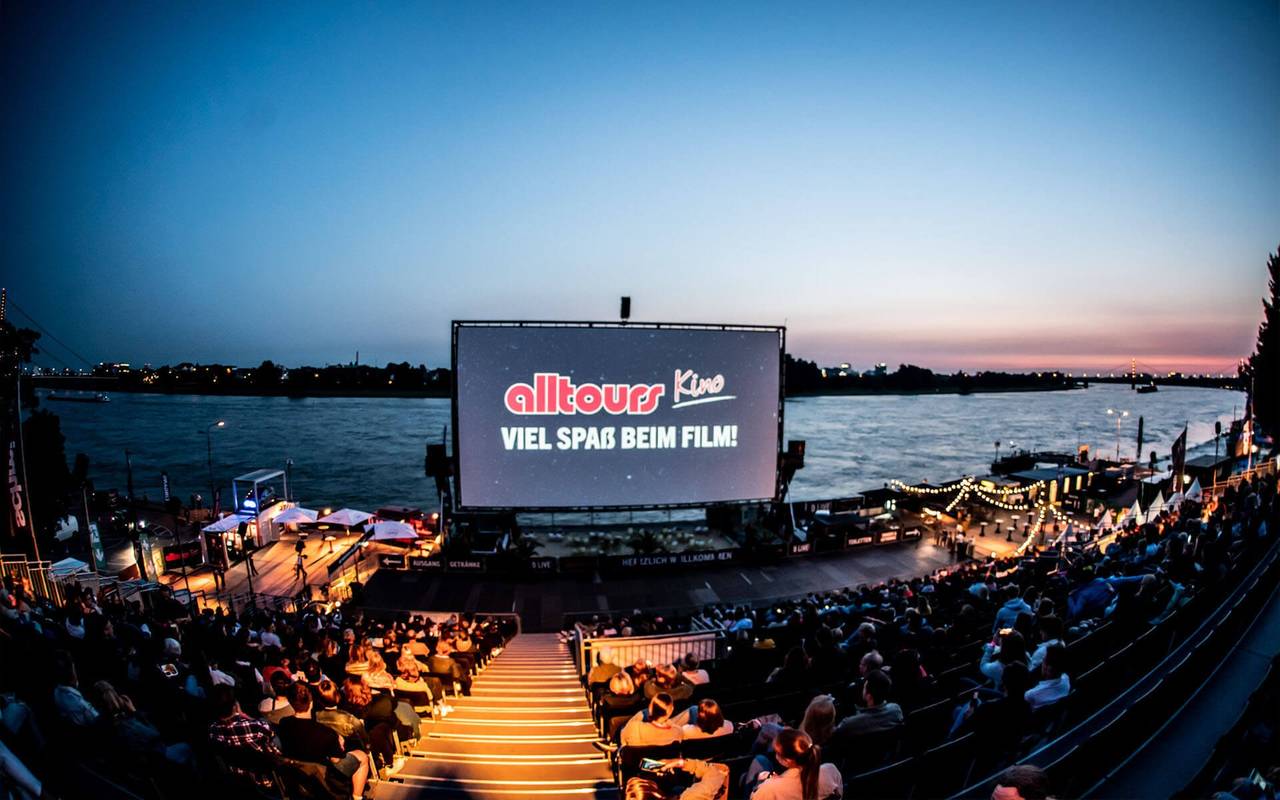 Zu sehen ist die Leinwand des alltours Open Air Kinos. Auf der Leinwand sieht man das alltours Logo. Im Vordergrund sieht man die Tribüne des Kinos, auf der viele Menschen sitzen - im Hintergrund sieht man den Rhein. Am Himmel geht grade die Sonne unter.