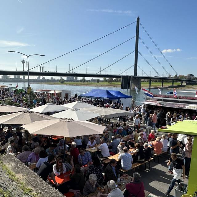 Blick auf den voll besuchten Fischmarkt am Düsseldorfer Rheinufer im Sonnenschein. Im Hintergrund ist die Oberkasseler Brücke und der Rhein zu sehen.