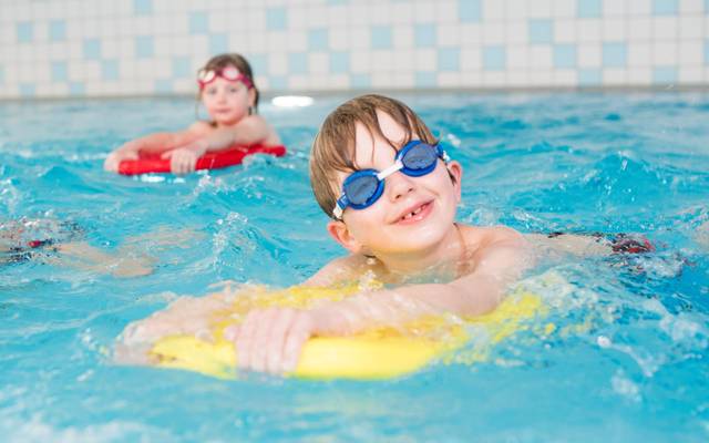 Ein Junge mit Schwimmbrille schaut in die Kamera. Er liegt auf einer gelben Schwimmhilfe.