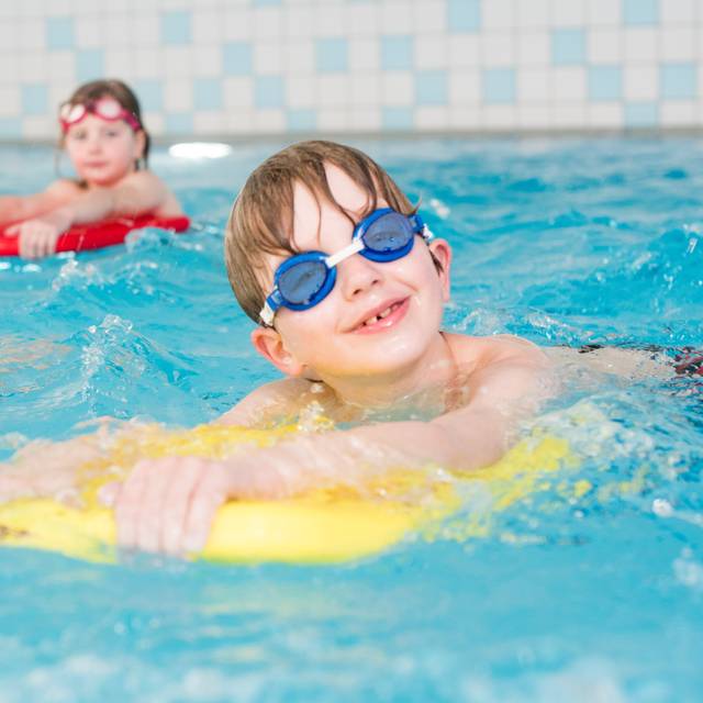 Ein Junge mit Schwimmbrille schaut in die Kamera. Er liegt auf einer gelben Schwimmhilfe.
