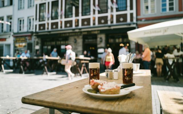 Im Vordergrund sind zwei Gläser Schlüssel-Alt und ein Teller mit einem Mettbrötchen zu sehen. Im Hintergrund die Fassade der Brauerei Zum Schlüssel.