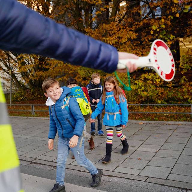 I-Dötzchen auf ihrem Schulweg. Ein Ordner regelt mit einer Kelle den Verkehr und sorgt so für Sicherheit.