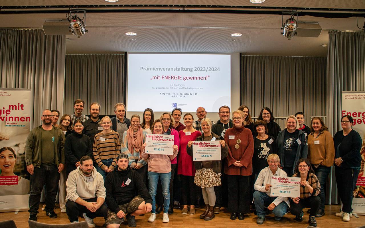 Gruppenfoto der teilnehmenden Einrichtungen des Programms "Mit Energie gewinnen!". Sie halten die überreichten Prämienschecks ins Bild. Im Hintergrund wird der Titel des Programms "Mit Energie gewinnen!" an die Wand projiziert.