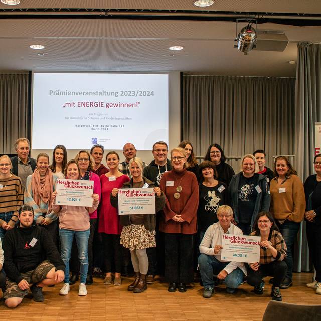 Gruppenfoto der teilnehmenden Einrichtungen des Programms "Mit Energie gewinnen!". Sie halten die überreichten Prämienschecks ins Bild. Im Hintergrund wird der Titel des Programms "Mit Energie gewinnen!" an die Wand projiziert.