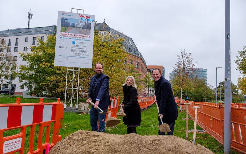 Der symbolische Spatenstich für die erste Radleitroute. OB Stephan Keller, Katharina Metzker Leiterin des Amtes für Verkehrsmanagement, und Michael Köhler, Geschäftsführer IPM Düsseldorf GmbH halten eine Schaufel in der Hand und stehen vor einem Sandberg.