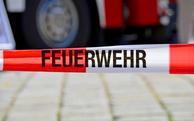 Ein rot-weißes Flatterband mit der Aufschrift "Feuerwehr". Im Hintergrund ist unscharf ein Feuerwehrwagen zu sehen.