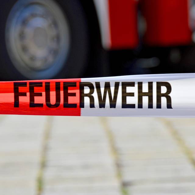 Ein rot-weißes Flatterband mit der Aufschrift "Feuerwehr". Im Hintergrund ist unscharf ein Feuerwehrwagen zu sehen.