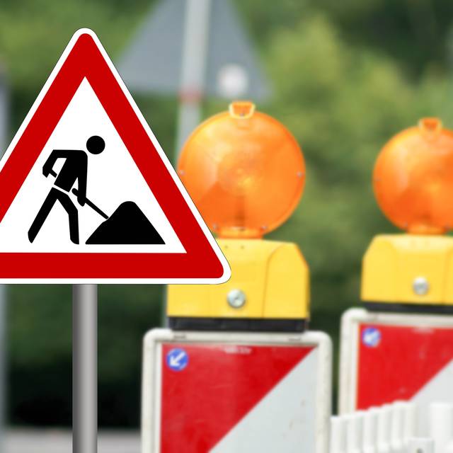 Ein Baustellenschild weist auf eine Baustelle hin. Im Hintergrund stehen zwei Warnbaken.