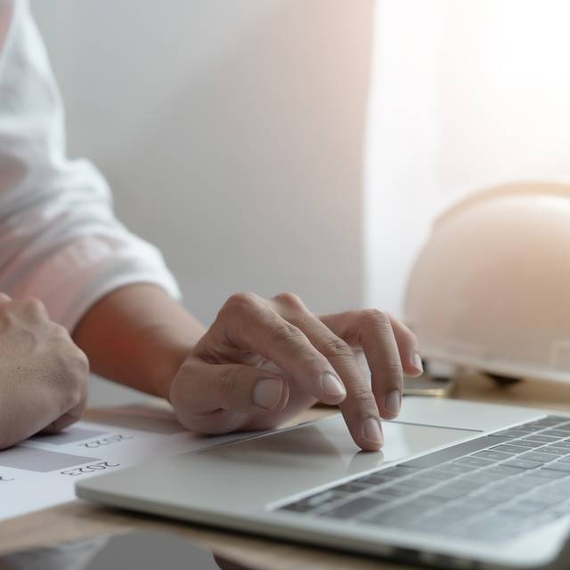 Eine Person tippt auf einem Laptop-Computer, während ein Bauhelm auf dem Schreibtisch liegt.