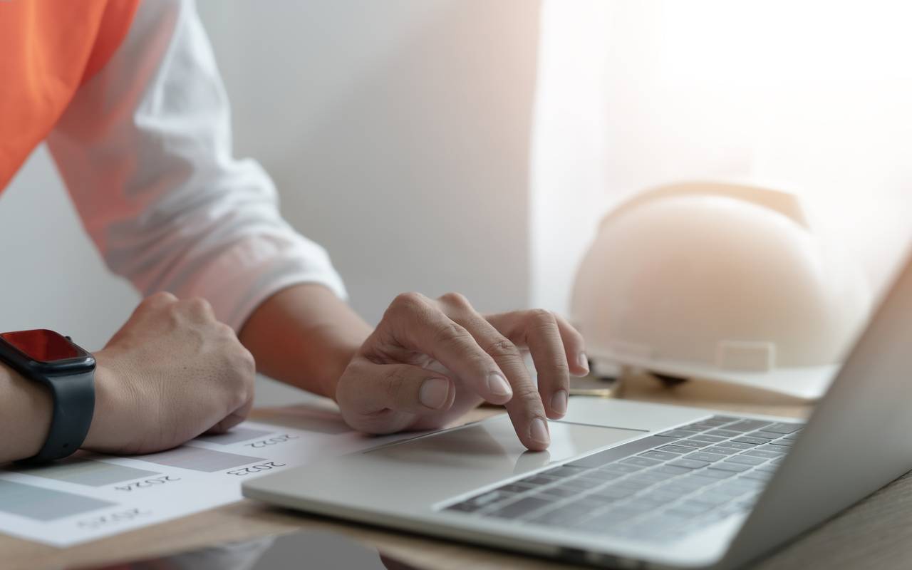 Eine Person tippt auf einem Laptop-Computer, während ein Bauhelm auf dem Schreibtisch liegt.