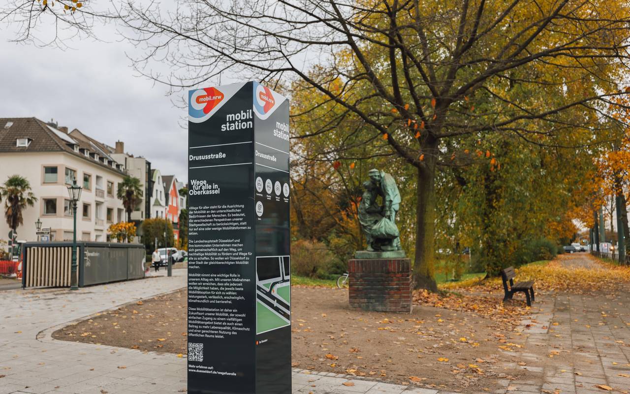 Eine Infostele weist auf die neue Mobilitätsstation an der Drususstraße in Oberkassel hin. Im Hintergrund sind Fahrradboxen zu erkennen.