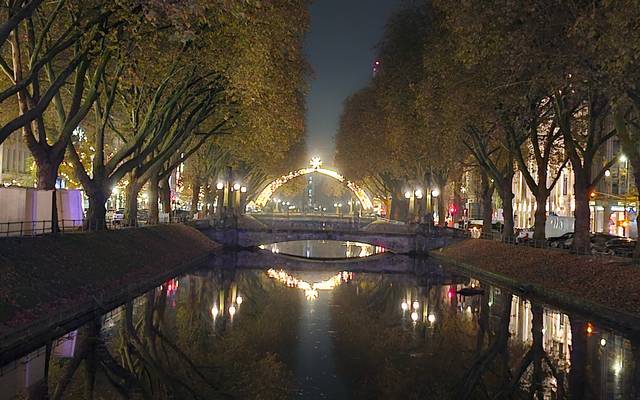 Die Königsallee mit dem Kö-Graben bei Nacht. Im Hintergrund leuchtet der Lichterdom auf der Steinstraße.