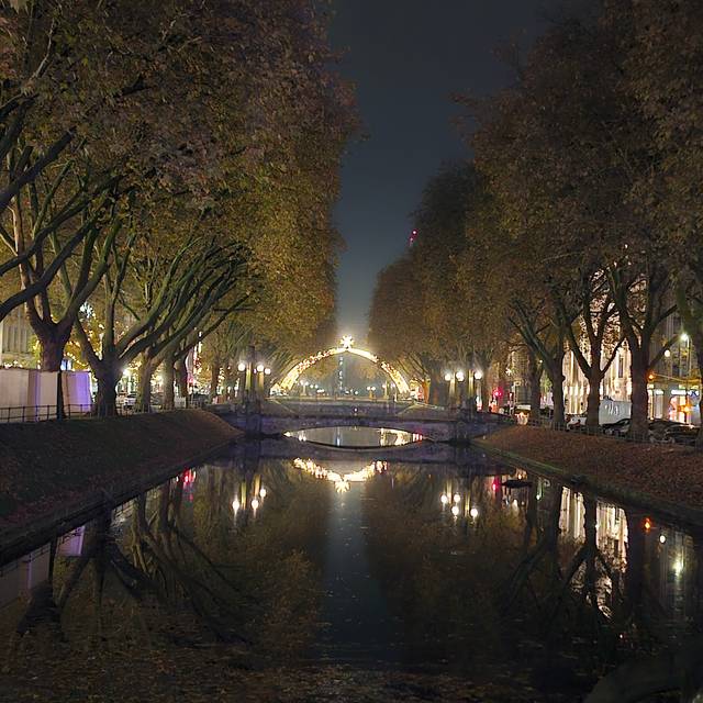 Die Königsallee mit dem Kö-Graben bei Nacht. Im Hintergrund leuchtet der Lichterdom auf der Steinstraße.
