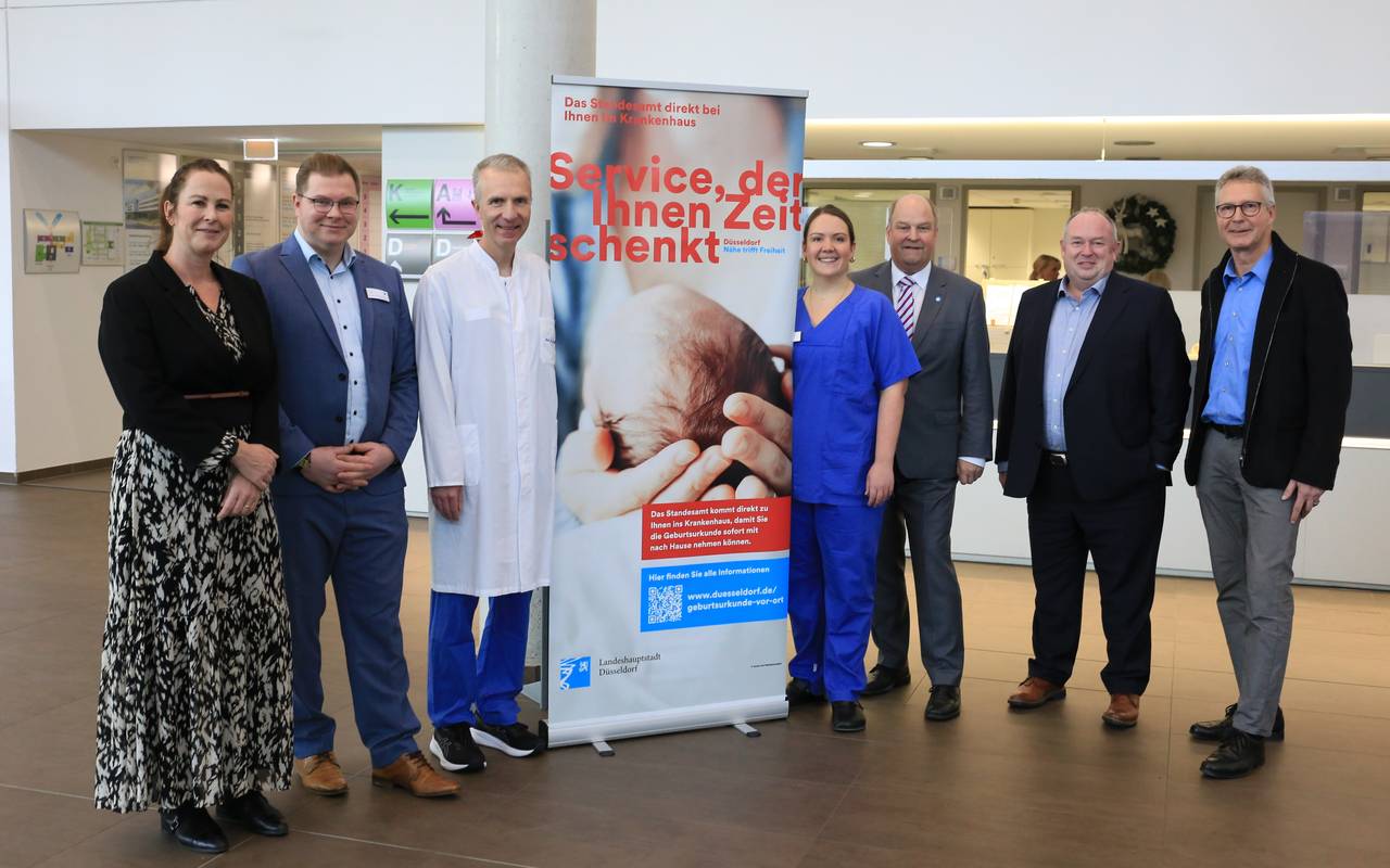Die schnelle Geburtsurkunde in der Düsseldorfer Diakonie präsentieren Annika Stahn (Standesamt), Dag Bill (Patientenverwaltung), Prof. Dr. med. Ralf Schmitz (Chefarzt), Isabel Meurer (Hebamme), Dr. Holger Stiller (Krankenhausdirektor), Olaf Wagner (Beigeordneter) und Harald Wehle (Amt für Einwohnerwesen). Sie stehen von links nach rechts neben einem Plakat mit weiteren Informationen.