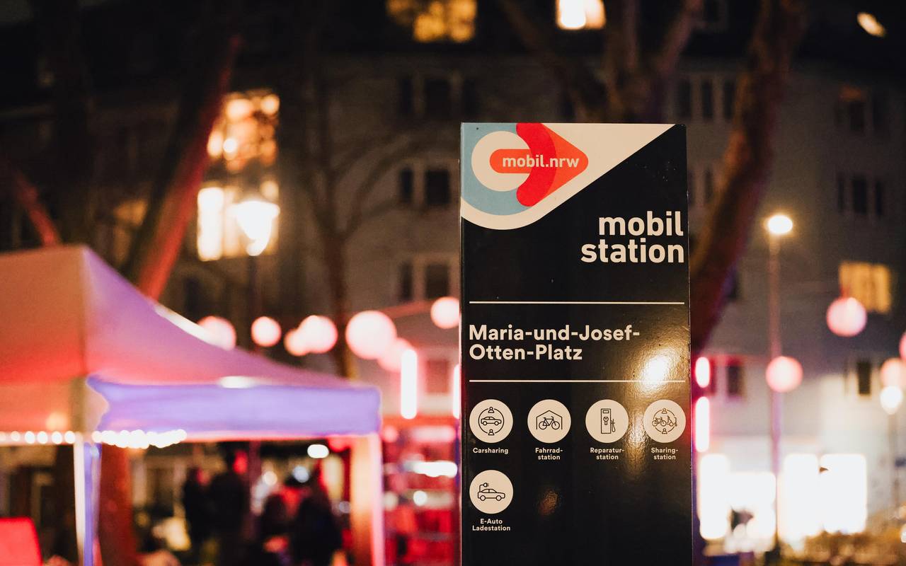 Eröffnung der neuen Mobilitätsstation am Maria-und-Josef-Otten-Platz in Pempelfort. Ein großes Schild weist auf die Angebote hin. Im Hintergrund sehen wir Zelte für die Aktionen am Eröffnungstag und die winterliche Beleuchtung entlang der Wohnhäuser.