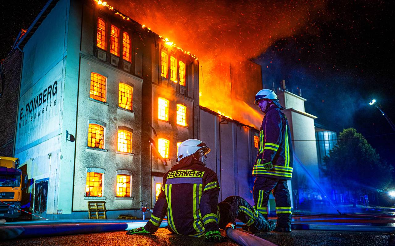 Zwei Feuerwehrleute vor einem brennenden Gebäude. Der eine sitzt erschöpft der andere steht.