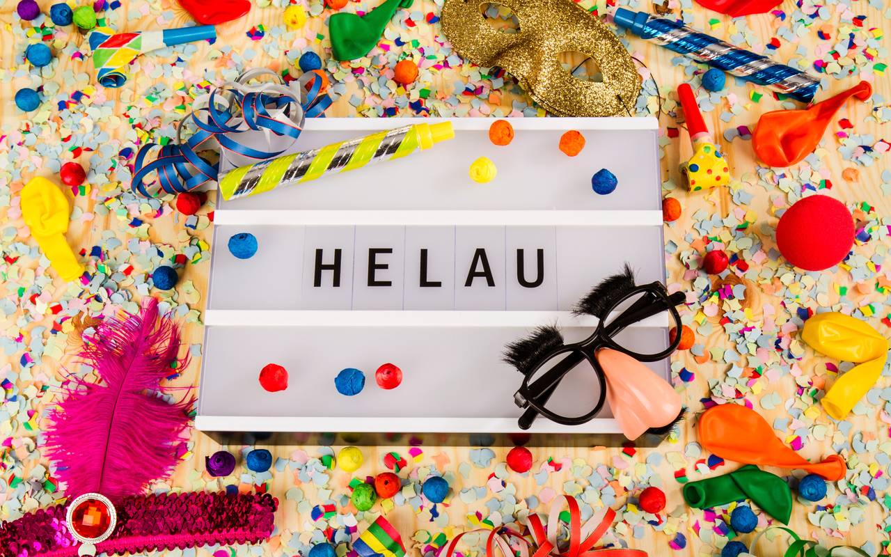 Ein festliches Szenario mit Konfetti, Luftballons, Partytröten, einer goldenen Maske und Spaßbrillen umgibt eine Leuchtbox mit der Aufschrift "HELAU."