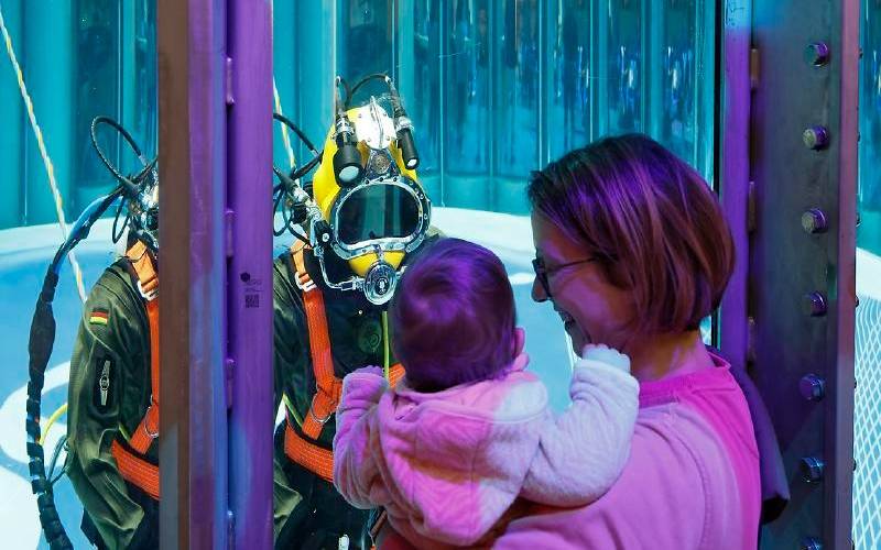 Eine Frau schaut mit ihrem Kind auf dem Arm auf einen Taucher, der hinter einer Scheibe im Tauchbecken ist.