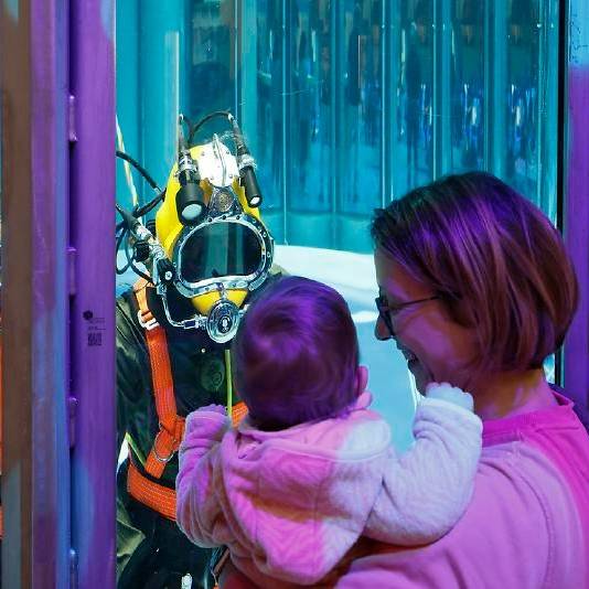 Eine Frau schaut mit ihrem Kind auf dem Arm auf einen Taucher, der hinter einer Scheibe im Tauchbecken ist.