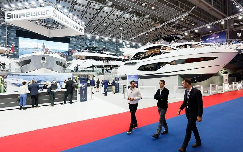 Messe boot: Drei Männer laufen im Vordergrund auf einem blau-roten Teppich durch die Messehalle. Sie unterhalten sich. Im Hintergrund kann man große Yachten und weiteres Publikum erkennen.