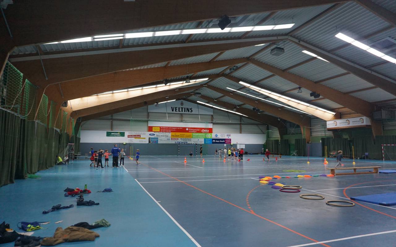 Zu sehen ist die Mehrzwecksporthalle in Düsseldorf Angermund. Im Hintergrund sieht man spielende Kinder, die an einem Training teilnehmen.