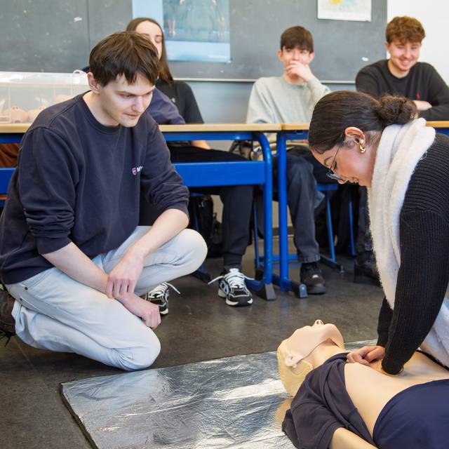 Eine Schülerin und ein Schüler üben die Wiederbelebung an einer Puppe. Im Hintergrund sitzen weitere Schüler.