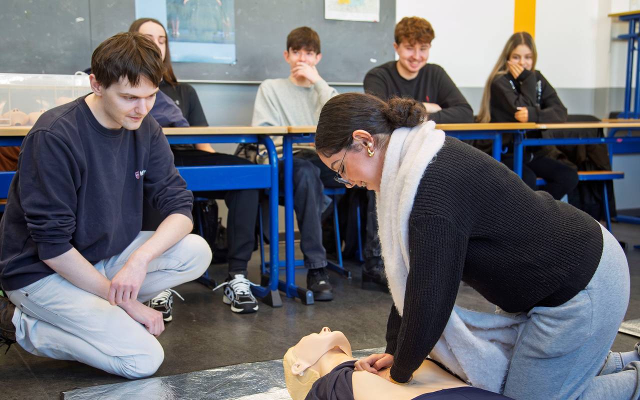 Eine Schülerin und ein Schüler üben die Wiederbelebung an einer Puppe. Im Hintergrund sitzen weitere Schüler.