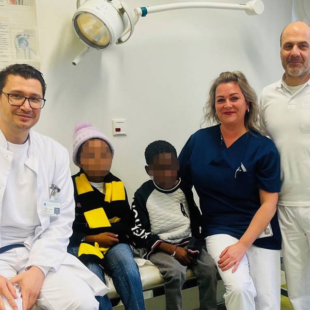 Zwei Männer und eine Frau stehen neben zwei Kindern, die auf einer Liege sitzen und deren Gesicher verpixelt sind