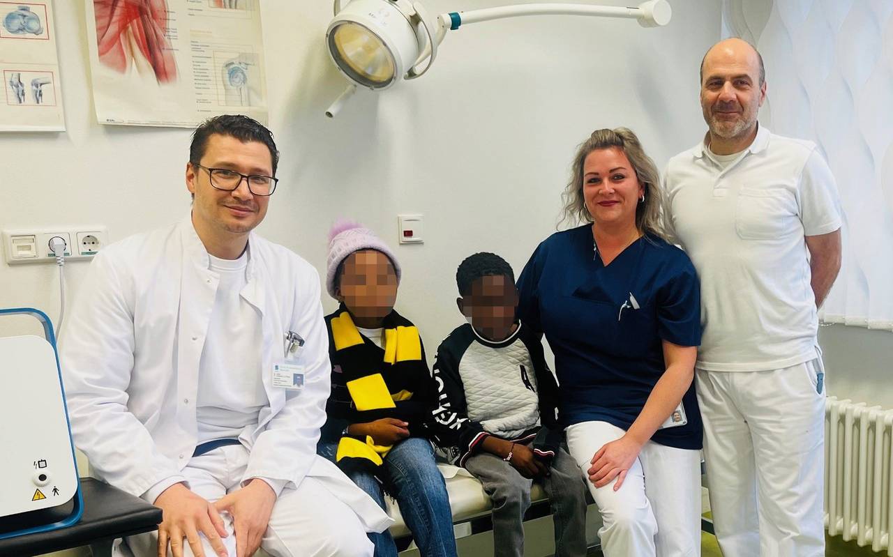 Zwei Männer und eine Frau stehen neben zwei Kindern, die auf einer Liege sitzen und deren Gesicher verpixelt sind