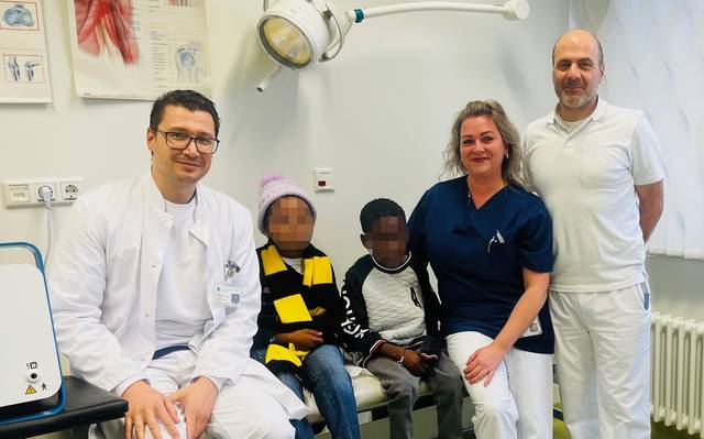 Zwei Männer und eine Frau stehen neben zwei Kindern, die auf einer Liege sitzen und deren Gesicher verpixelt sind