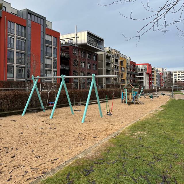 Eine Spielplatz, bestehend aus einer Sandfläche mit Spielgeräten. Im Vordergrund sind Schaukeln. Hinter dem Spielplatz stehen Mehrfamilienhäuser.