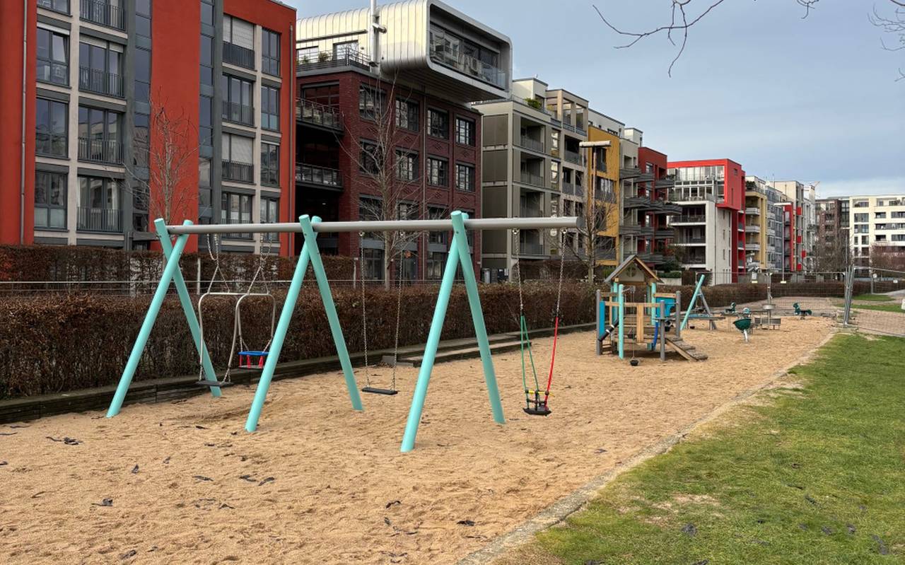 Eine Spielplatz, bestehend aus einer Sandfläche mit Spielgeräten. Im Vordergrund sind Schaukeln. Hinter dem Spielplatz stehen Mehrfamilienhäuser.