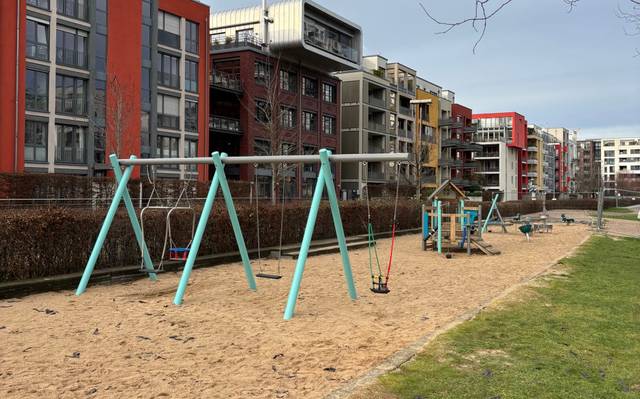 Eine Spielplatz, bestehend aus einer Sandfläche mit Spielgeräten. Im Vordergrund sind Schaukeln. Hinter dem Spielplatz stehen Mehrfamilienhäuser.
