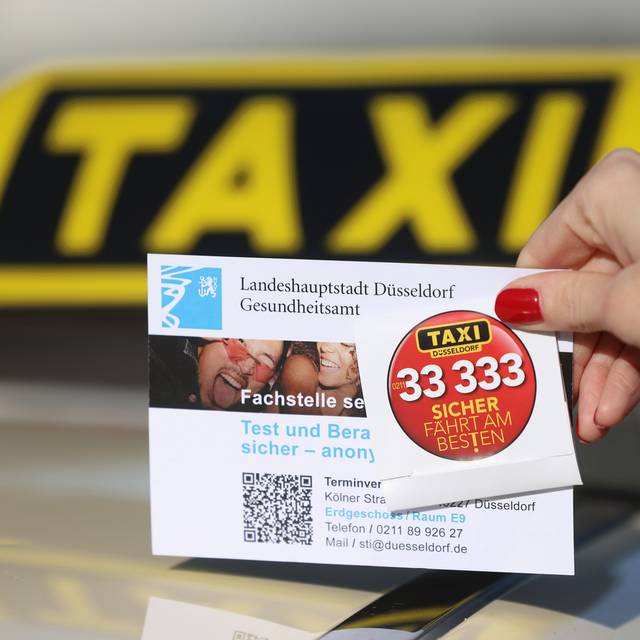 Foto zur Kondomaktion der Taxi-Innung Düsseldorf. Eine Frau hält ein Werbeschild mit Kondom vor einem Taxi-Schild.
